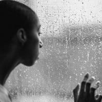 A man being depressed looking out the window to the rain outside.